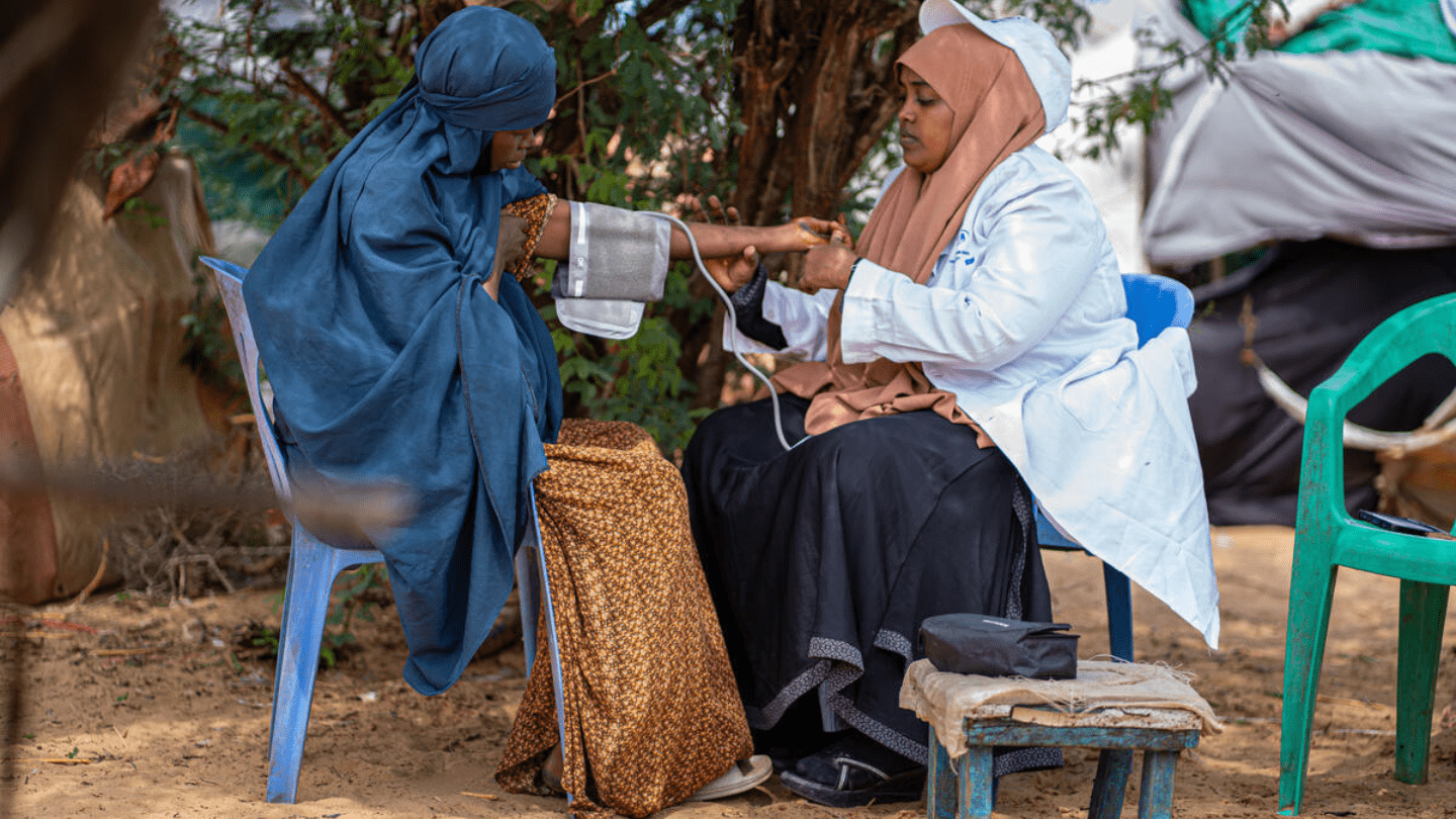 A midwifw in a white lab coat and hijab is taking the blood pressure of a woman seated in a blue plastic chair outdoors. Both women are wearing traditional clothing and head coverings. The setting is a makeshift medical area with simple seating and draped fabric in the background