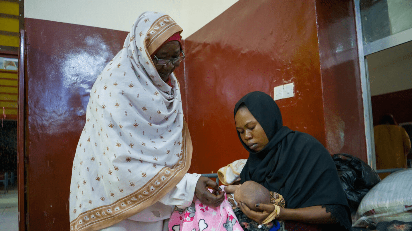 "Two women are in an indoor setting. The woman on the left, wearing a light-colored patterned head covering and a light-colored garment, is looking down and touching the hand of a newborn baby. The baby is being held by the woman on the right, who is wearing a dark head covering and a dark garment.