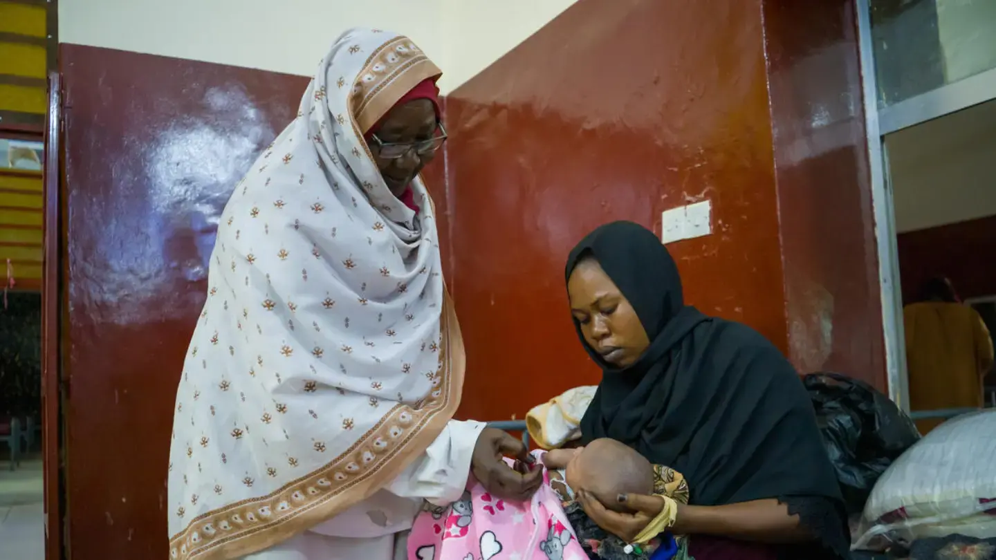 "Two women are in an indoor setting. The woman on the left, wearing a light-colored patterned head covering and a light-colored garment, is looking down and touching the hand of a newborn baby. The baby is being held by the woman on the right, who is wearing a dark head covering and a dark garment.